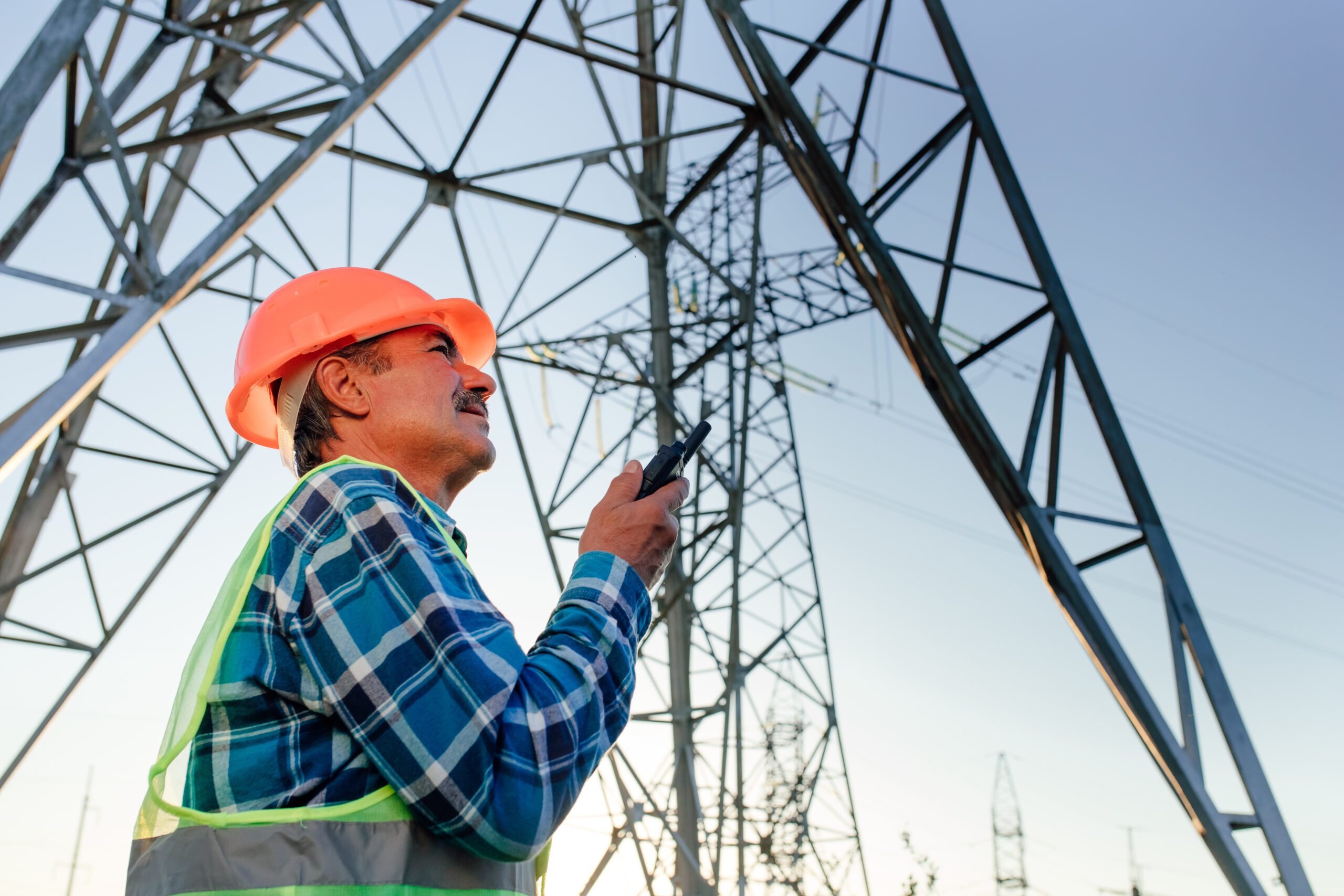 Low angle side view of middle aged male technician in hardhat speaking on walkie talkie while standing in back lit against electric power poles